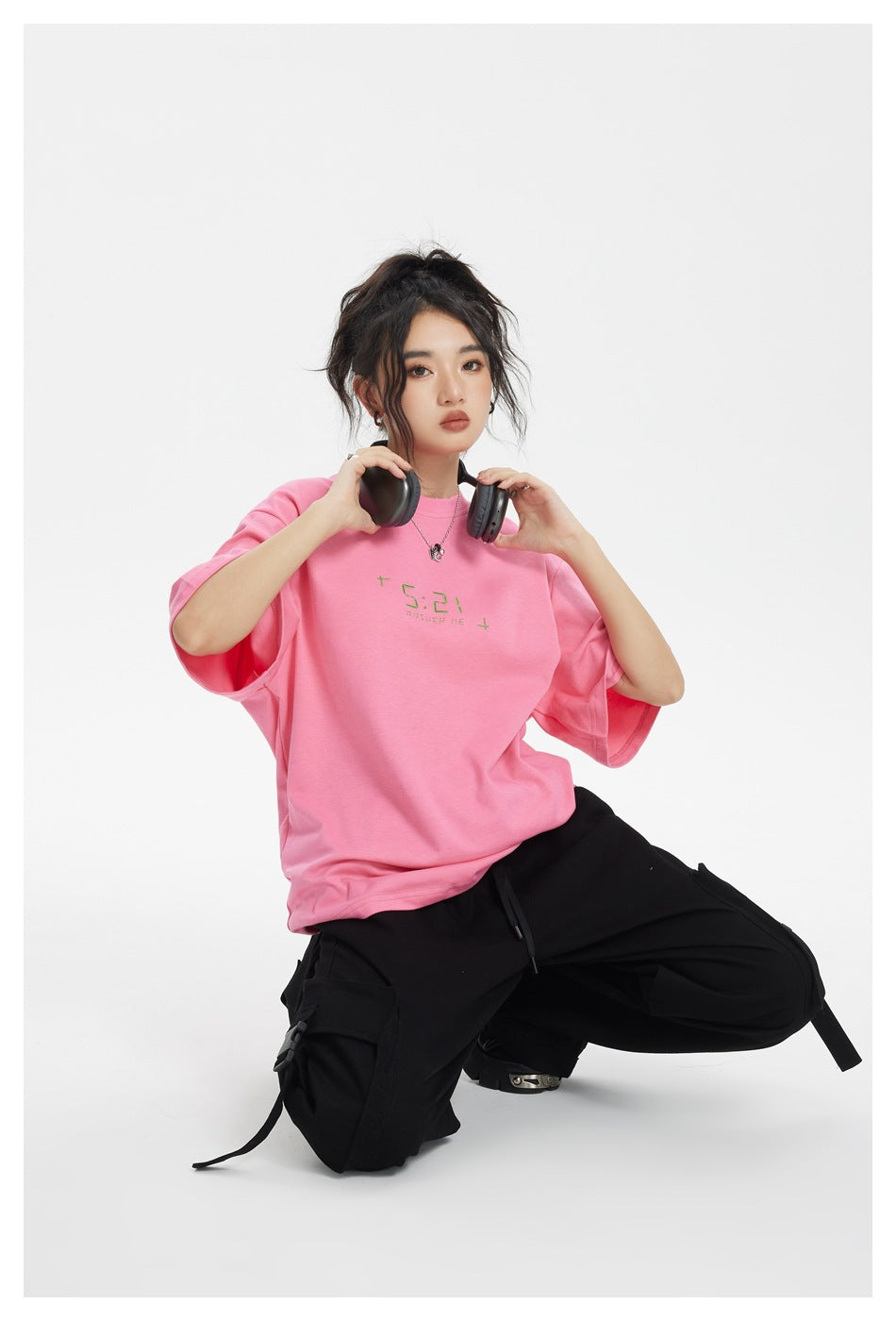 Model kneeling in a bright pink oversized T-shirt and black cargo pants, holding headphones at her shoulders.