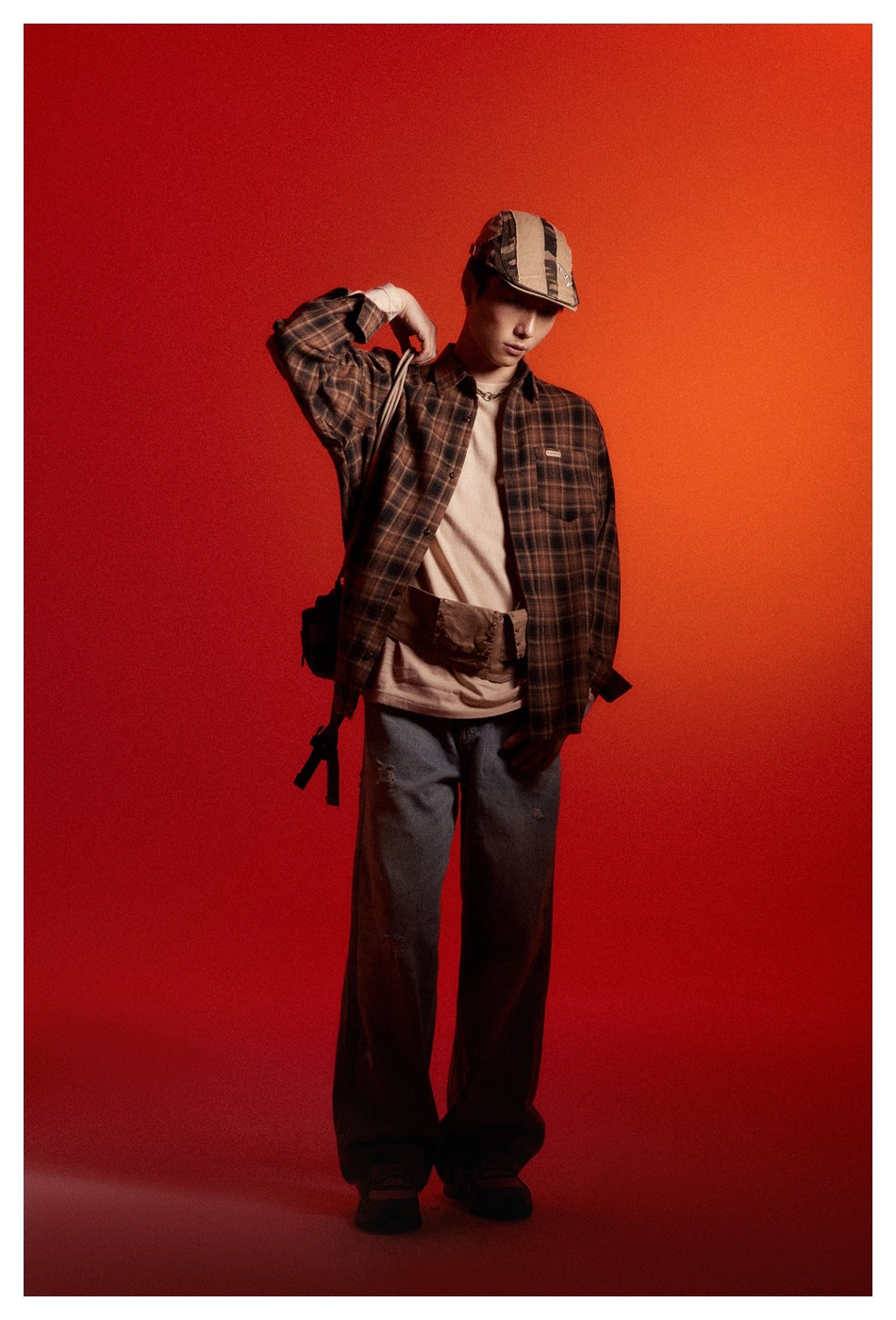 Model wearing an open dark brown plaid shirt over a beige top with loose dark jeans and a striped cap.