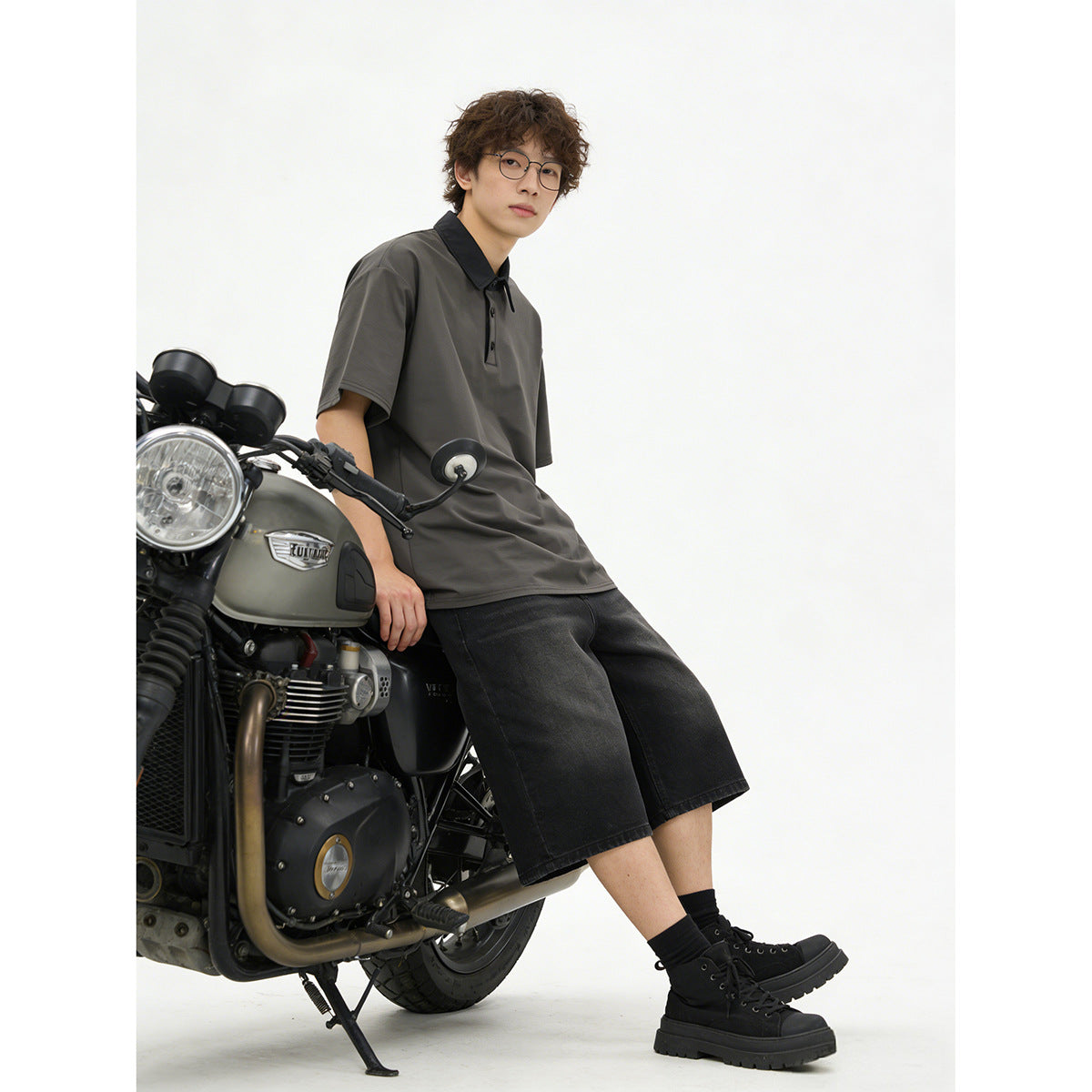 Model seated on a motorcycle wearing a charcoal polo with black collar, black denim shorts, and black boots.