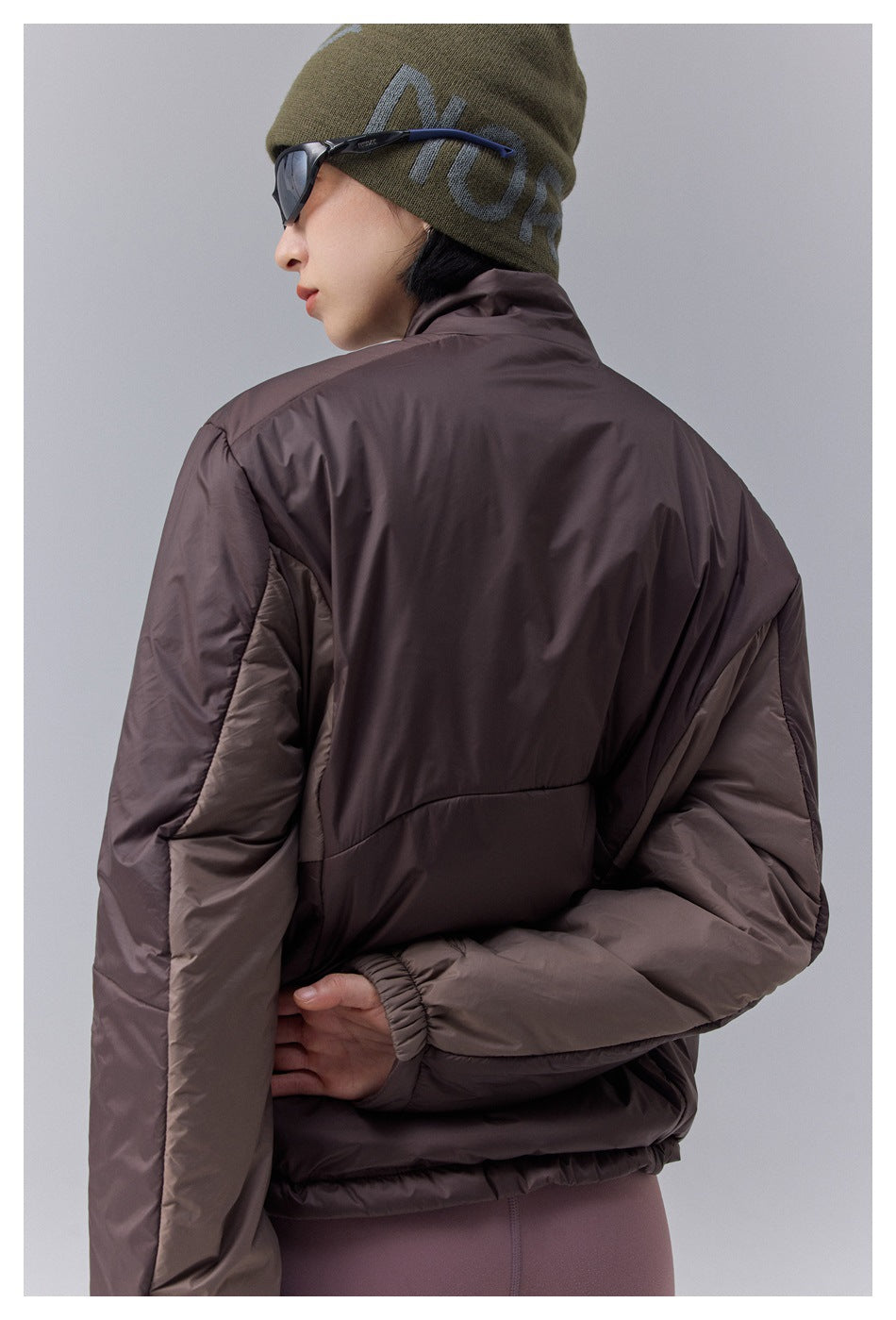 Rear view of a brown puffer jacket on a model, showing its color-blocked sleeves and elastic hem.