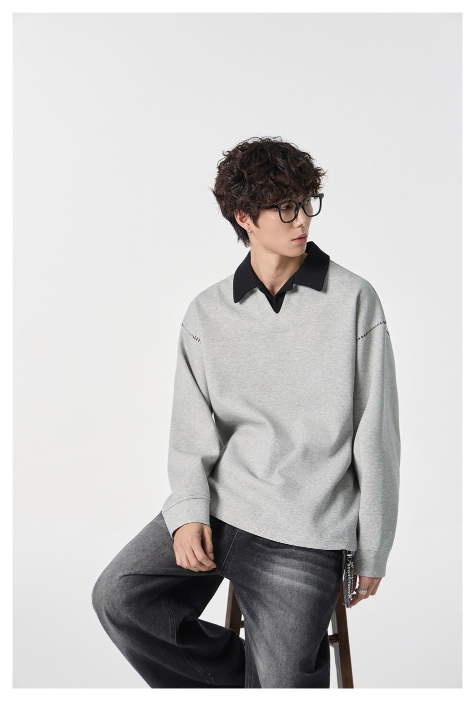 Seated model wearing a light gray pullover with a black collar and stitched shoulder details.
