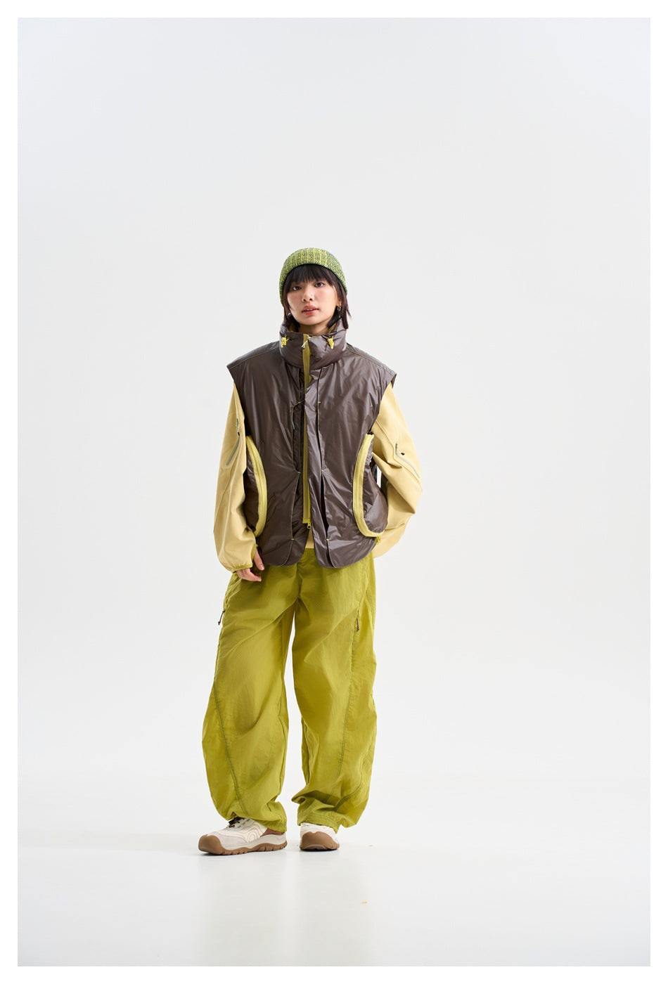Full-length model wearing a dark brown sleeveless vest over a pale yellow top, with lime trousers and a green beanie.