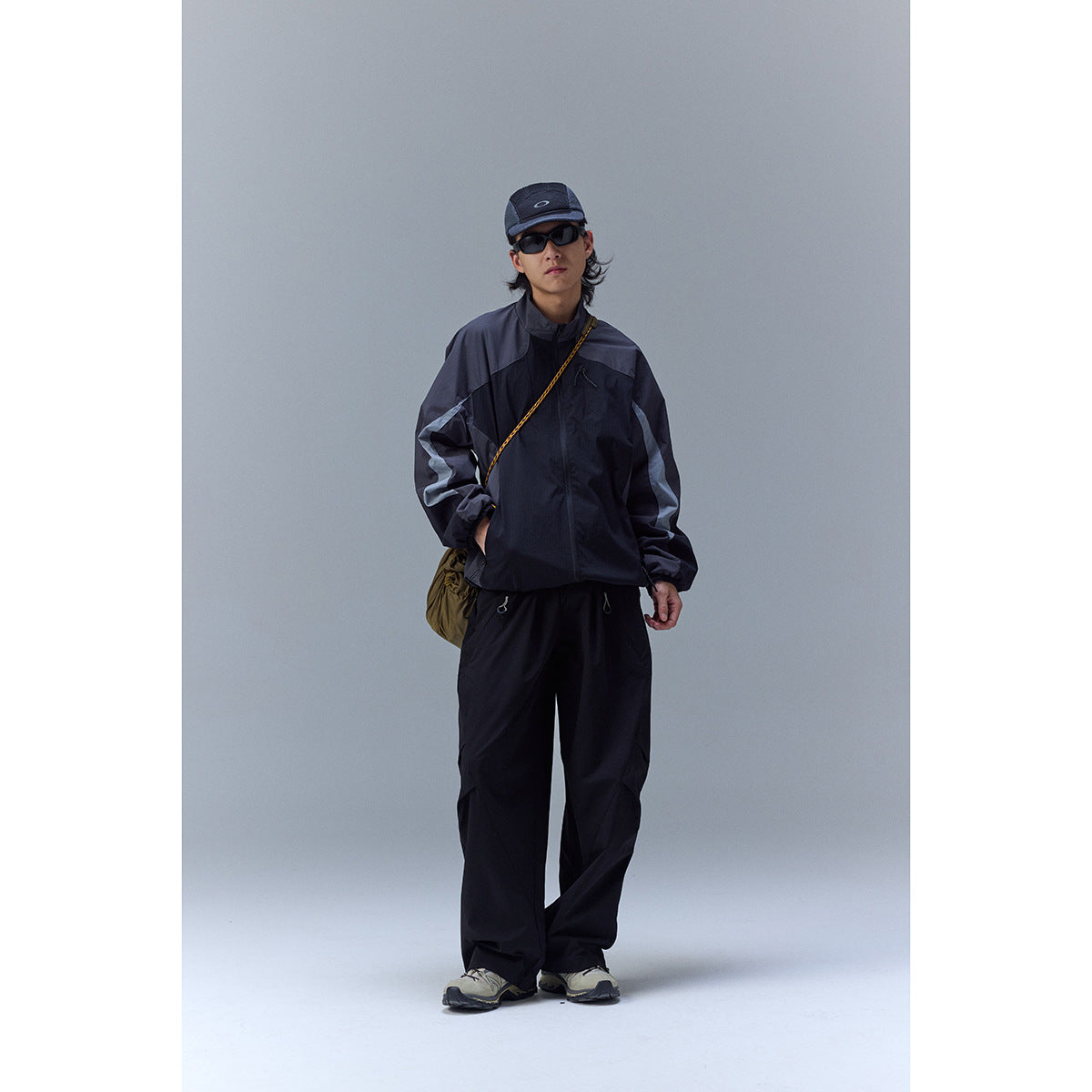 Model wearing a black and charcoal color-block jacket with matching loose pants, cap, and sunglasses.