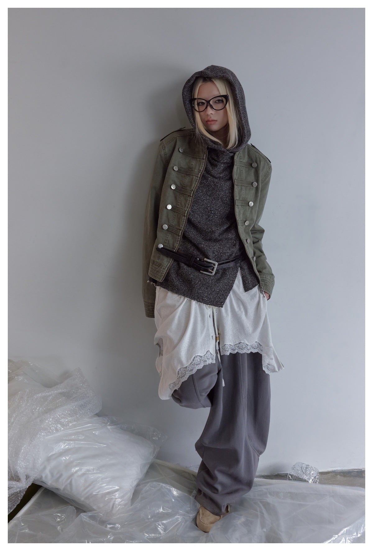 Model in an olive button-detailed jacket, charcoal hooded top, white lace-trim layer, and loose gray trousers.