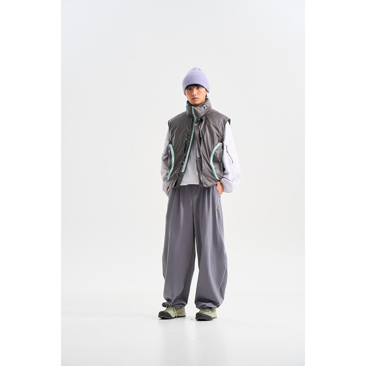 Model wearing a glossy charcoal sleeveless vest with aqua trim, white sleeves, loose gray trousers and a lavender beanie.