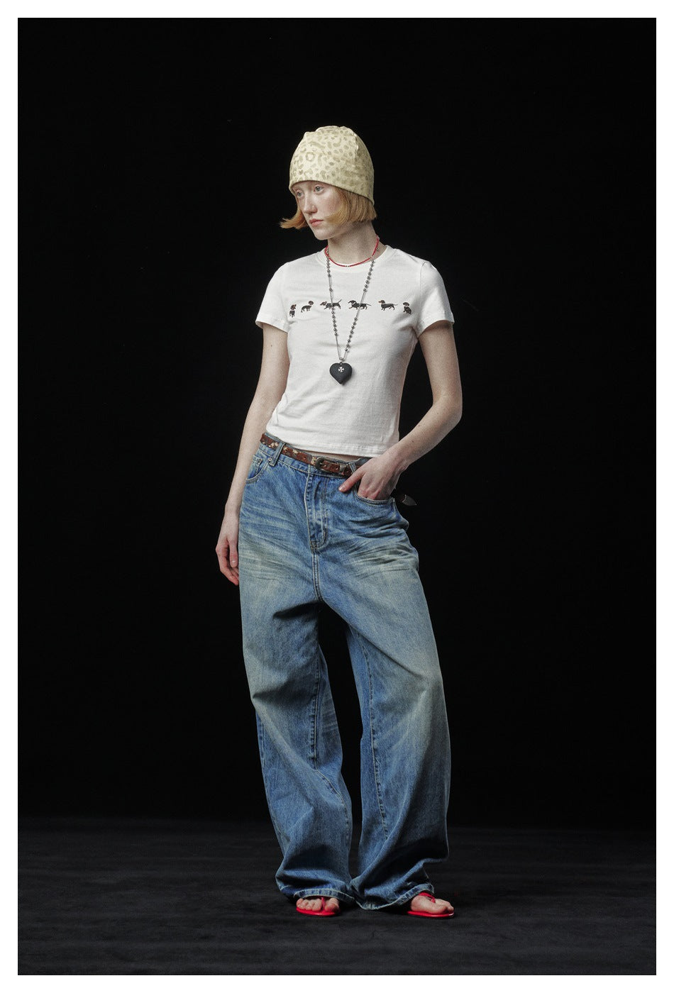 Model in a white dachshund-print T-shirt, loose faded blue jeans, a beanie and red sandals, seen from the side