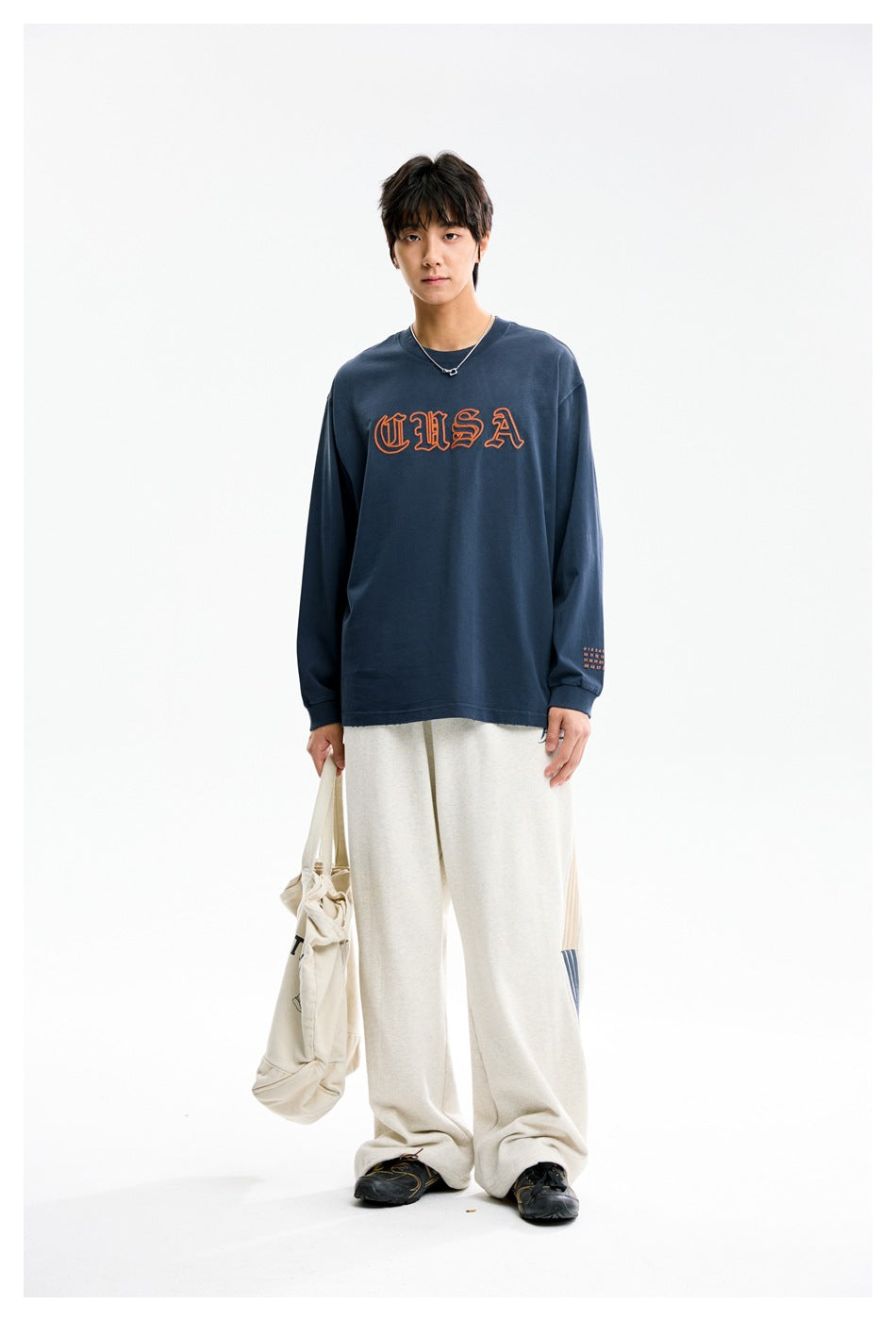 Model wearing a blue long-sleeve top with orange CUSA embroidery, light wide-leg pants and holding a canvas tote bag