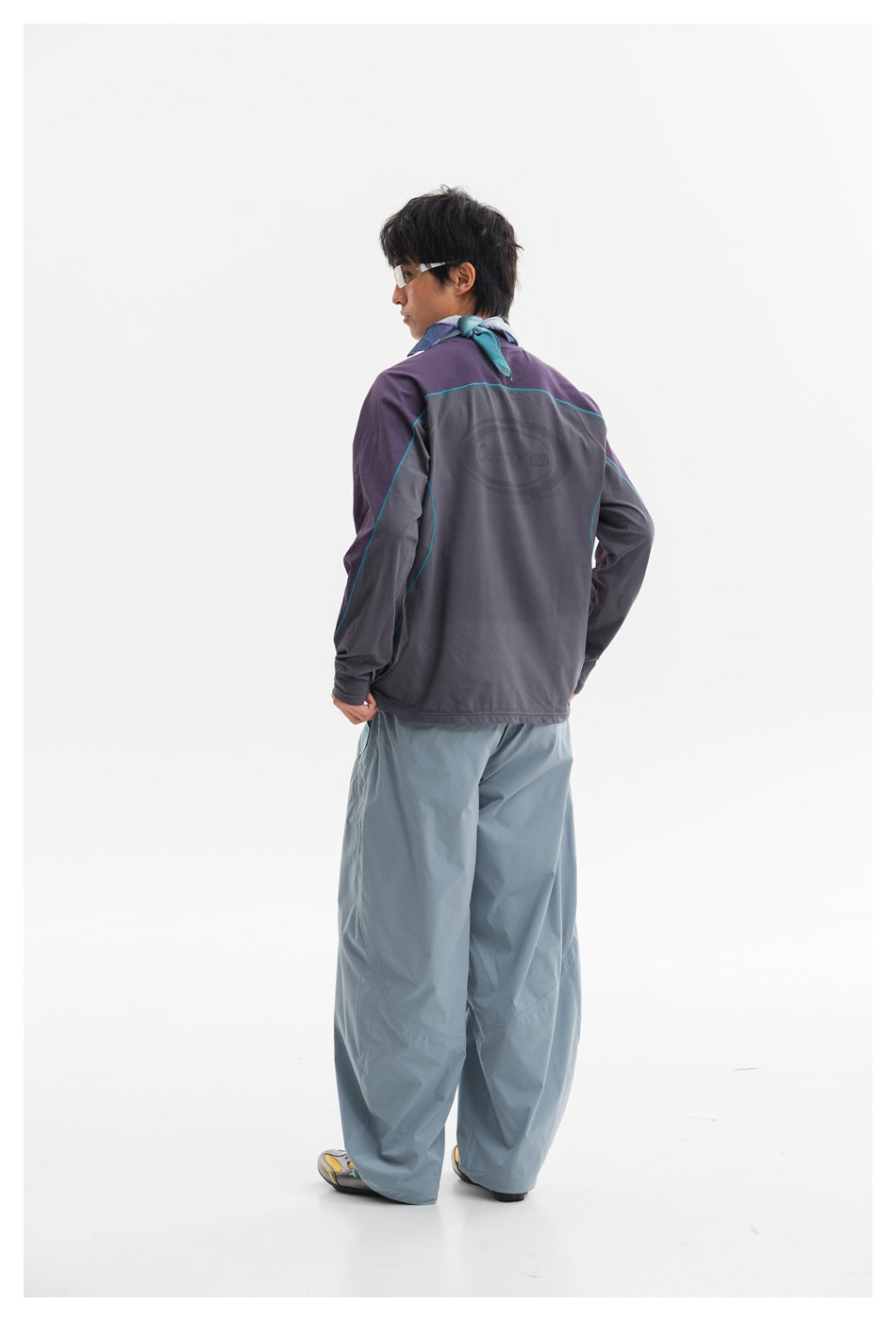 Back view of a model in a charcoal and purple long-sleeve top with teal piping and loose blue-gray trousers.