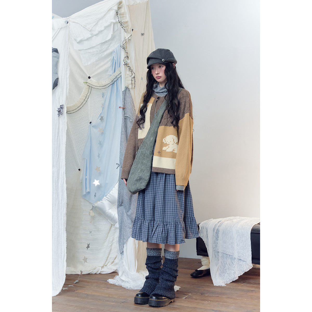 Model wearing a brown and mustard color-block cardigan, blue plaid tiered skirt, gray cap, and dark leg warmers.