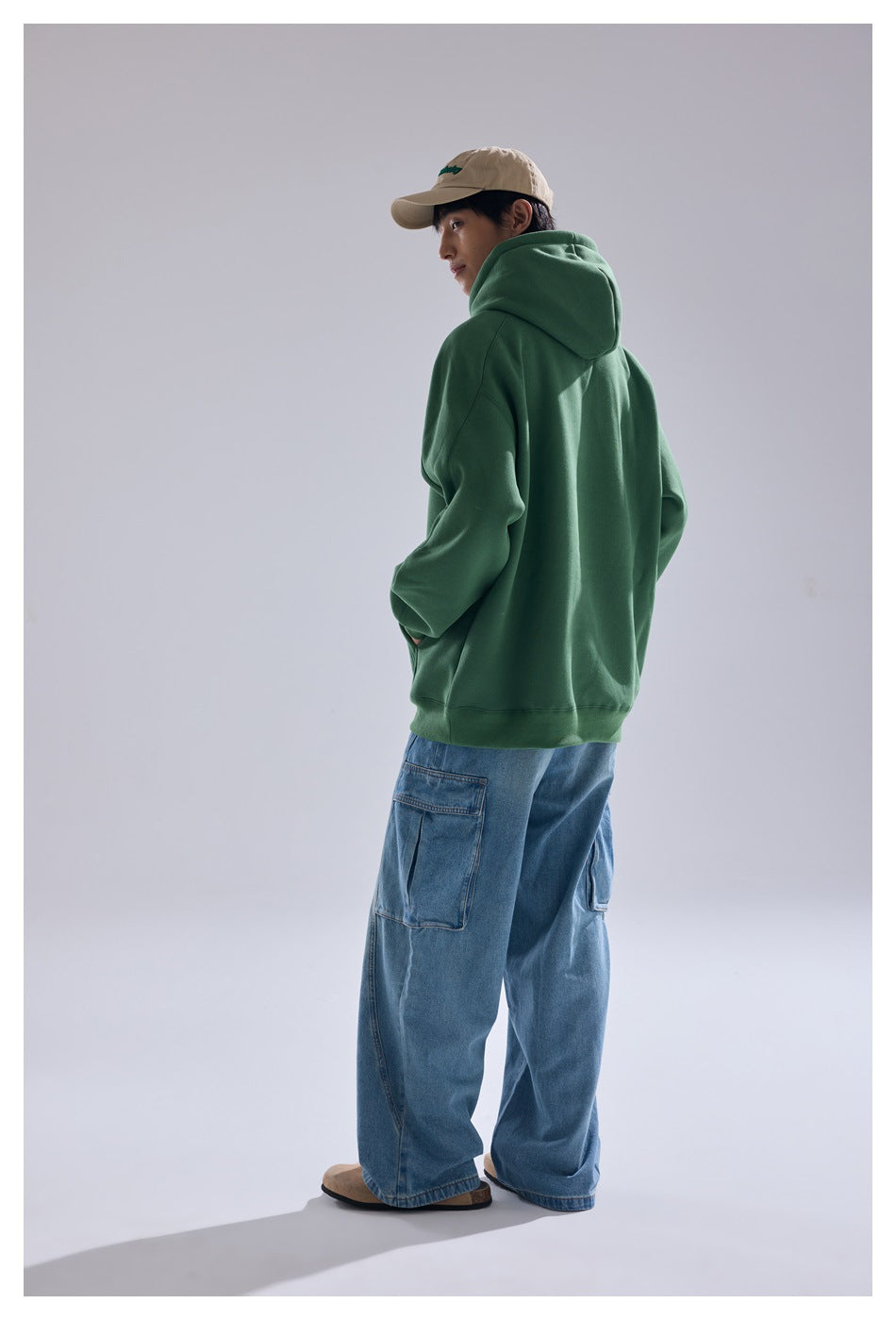 Model wearing a forest-green hoodie, loose blue cargo jeans, and a beige cap, shown mostly from behind.