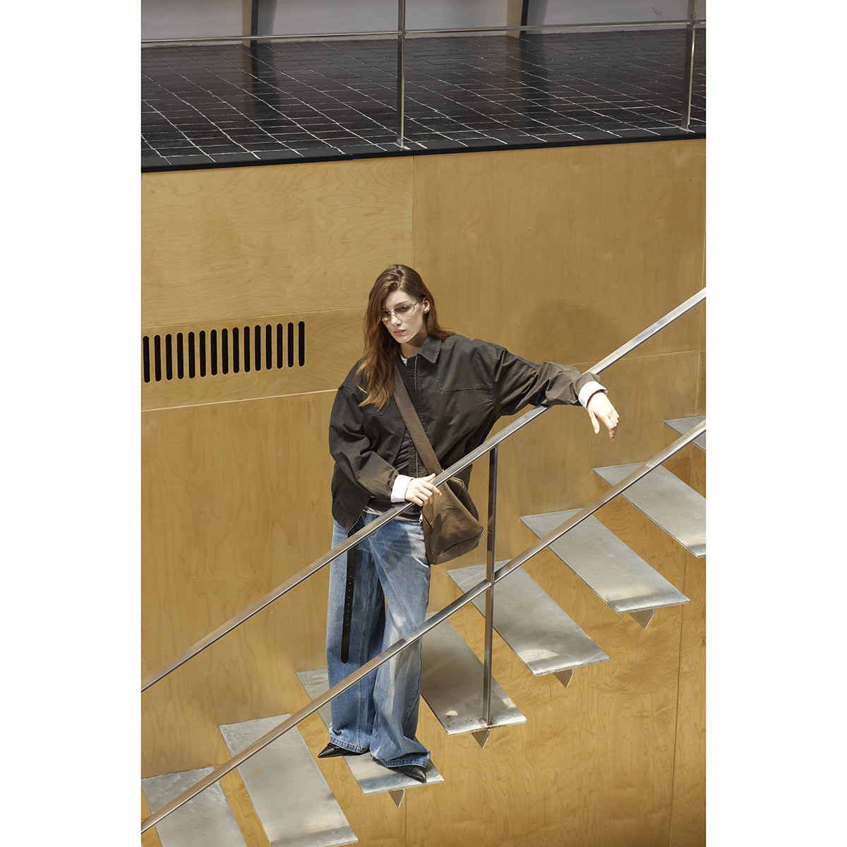 Model in a charcoal oversized jacket and wide-leg blue jeans on a modern staircase, carrying a brown shoulder bag.