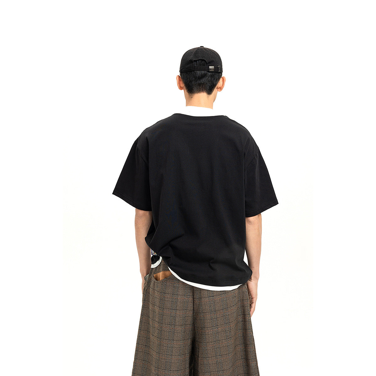 Rear view of a model wearing a loose black short-sleeve top over a white shirt, with plaid trousers.