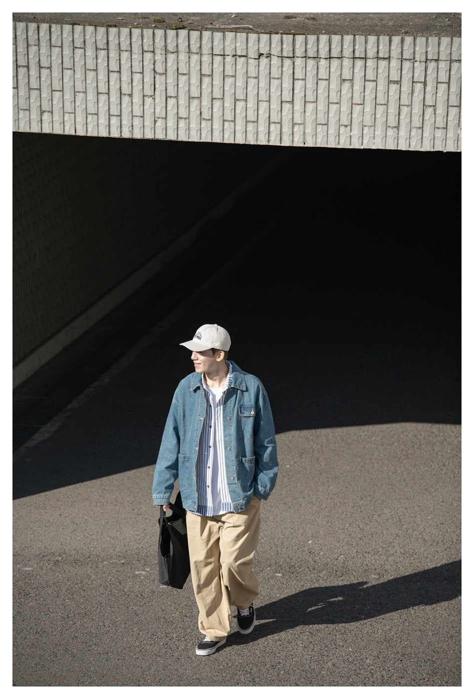 Full-length model wearing a blue denim jacket, striped shirt, tan trousers, white cap, and black sneakers outdoors.
