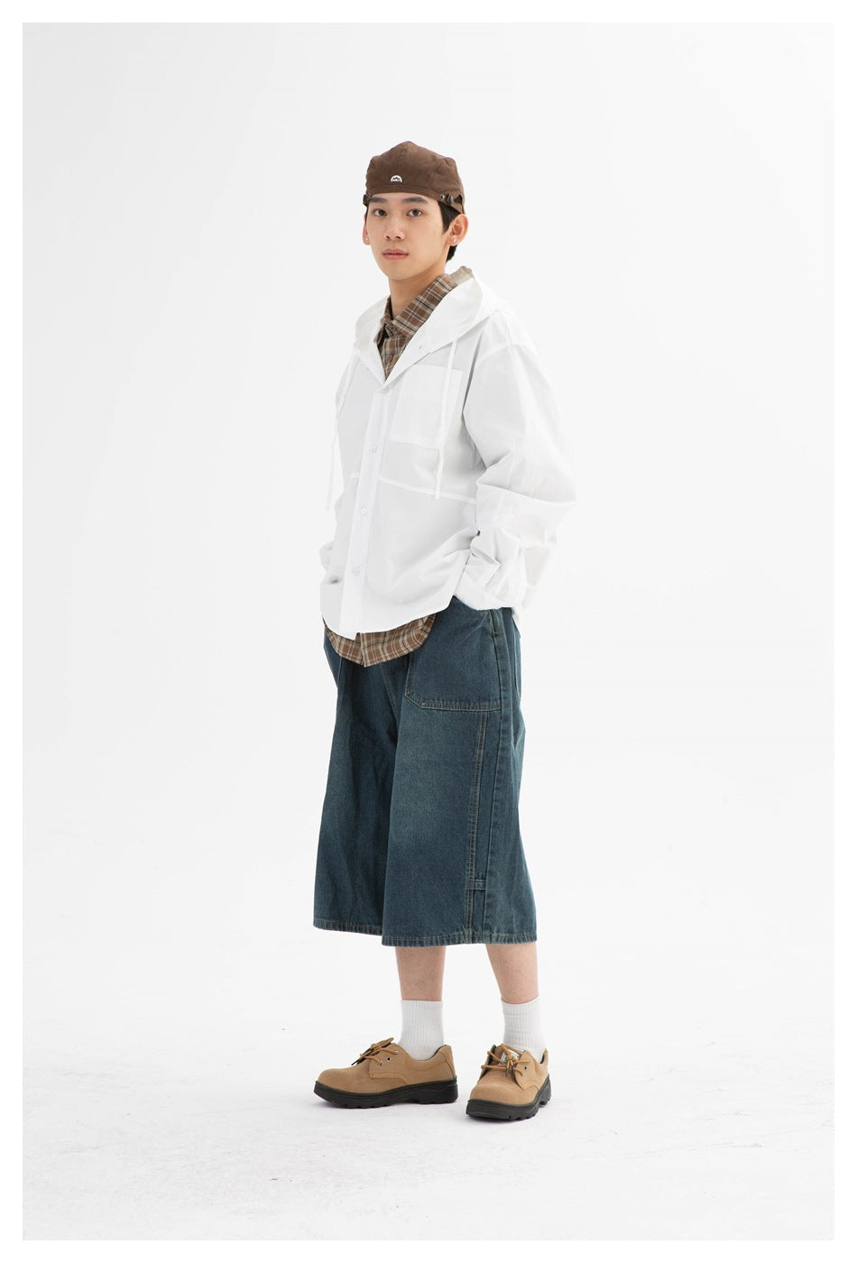 Full-length model wearing a white hooded button-front jacket over a brown plaid shirt and wide dark denim shorts.