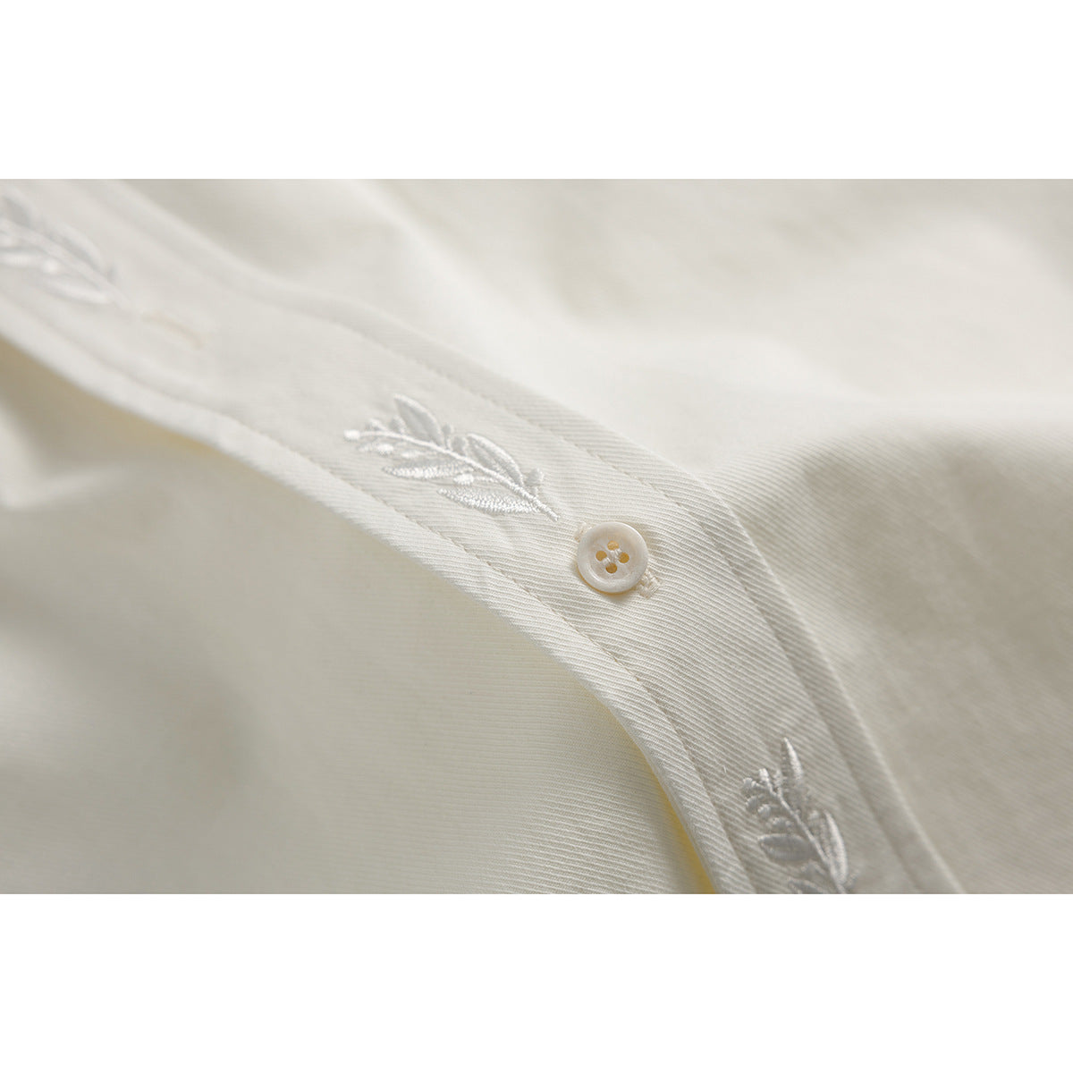 Close-up of an ivory shirt placket with white leaf embroidery and a pale four-hole button.