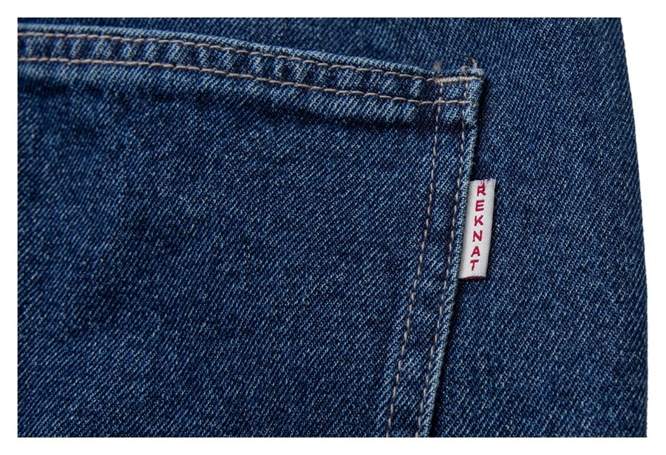 Close-up of a dark blue denim pocket edge with a small white REKNAT fabric tag.