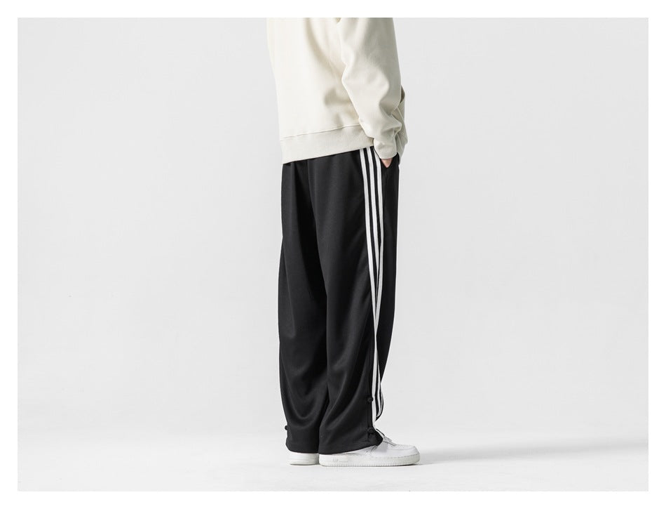 Rear view of a cropped model wearing loose black track pants with three white side stripes.
