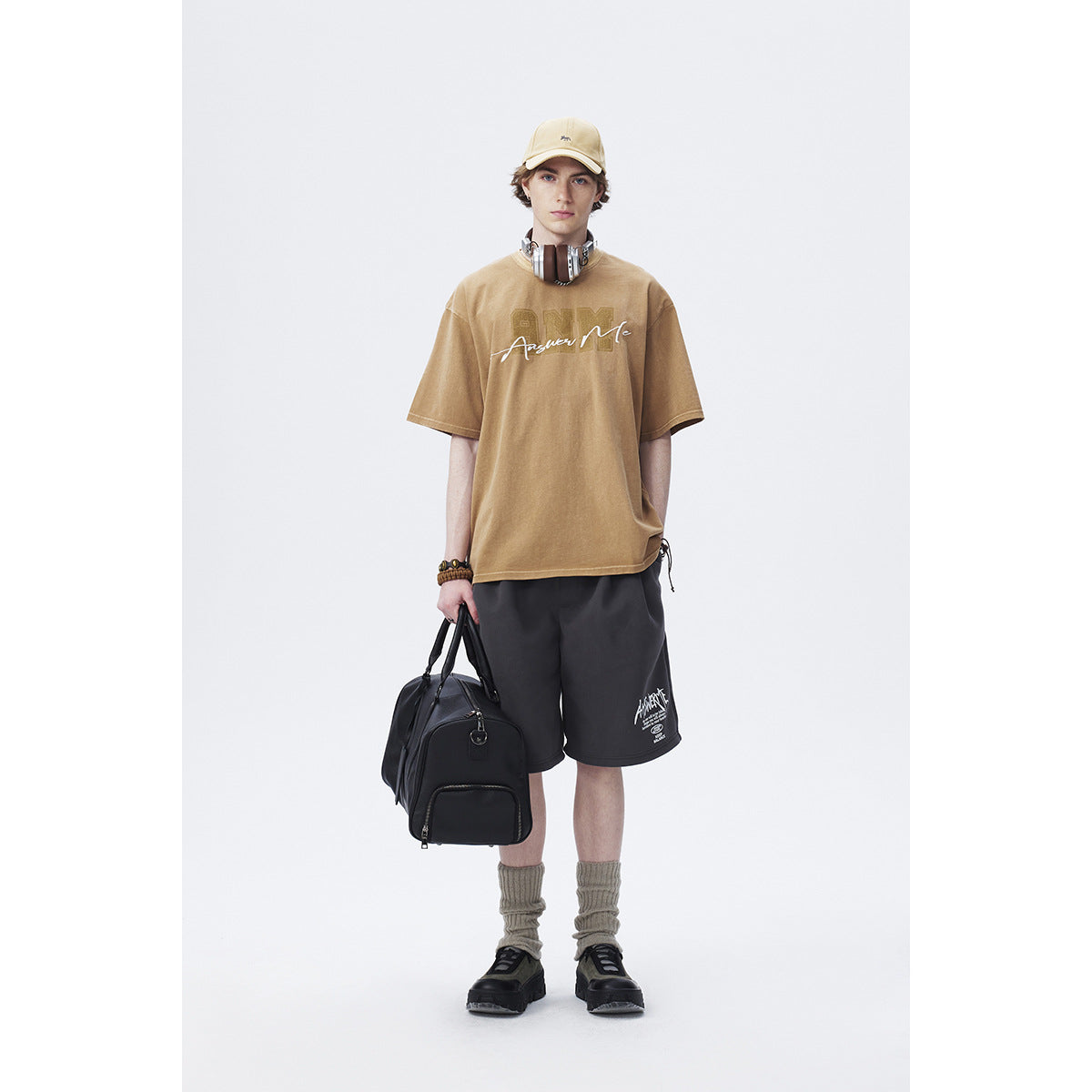 Model wearing a tan graphic T-shirt with black shorts, a beige cap, slouch socks and black shoes.