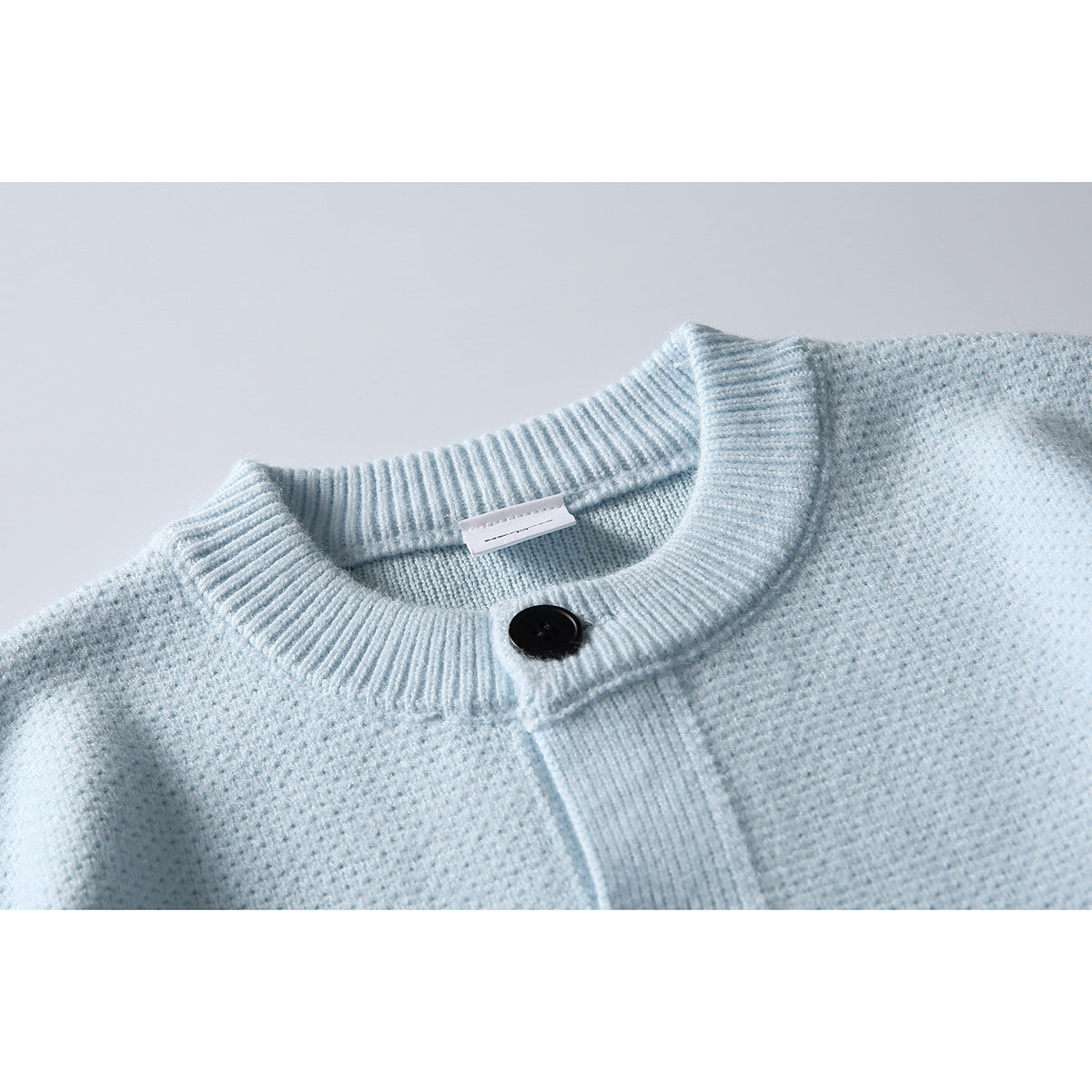 Close-up of a pale blue knitted cardigan showing its ribbed round neck, black button and front placket.