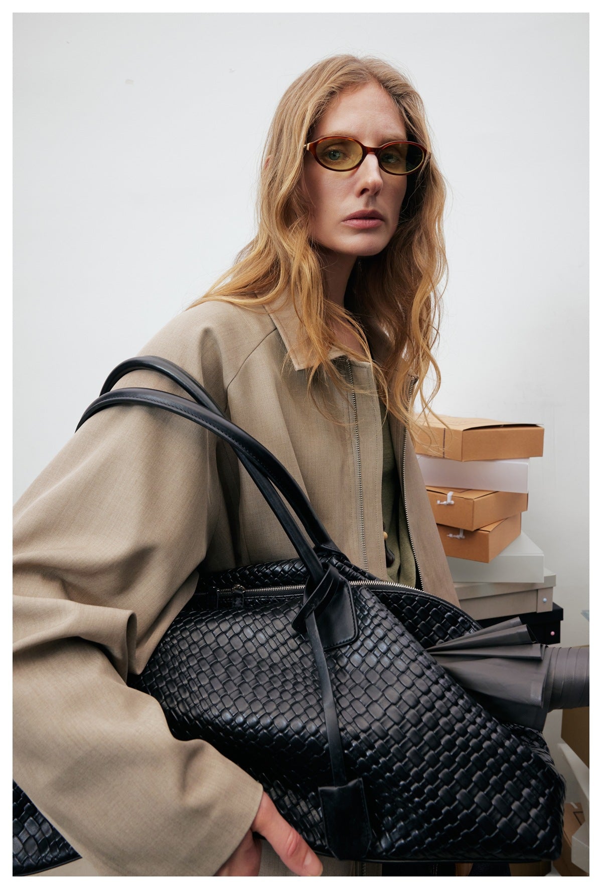 Close-up of a model in a taupe jacket carrying a large black woven handbag.