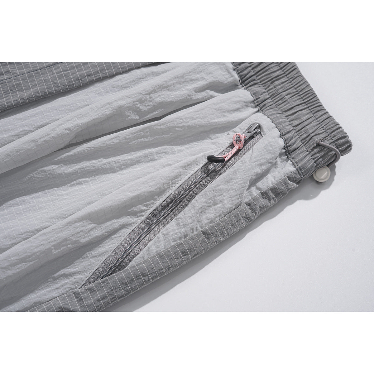 Close-up of gray pants showing a diagonal zip pocket with a pink pull, elastic waistband, and grid-patterned trim.