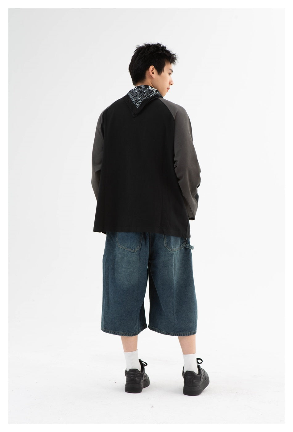 Back view of a model wearing a black sweatshirt with charcoal sleeves and loose blue denim shorts.