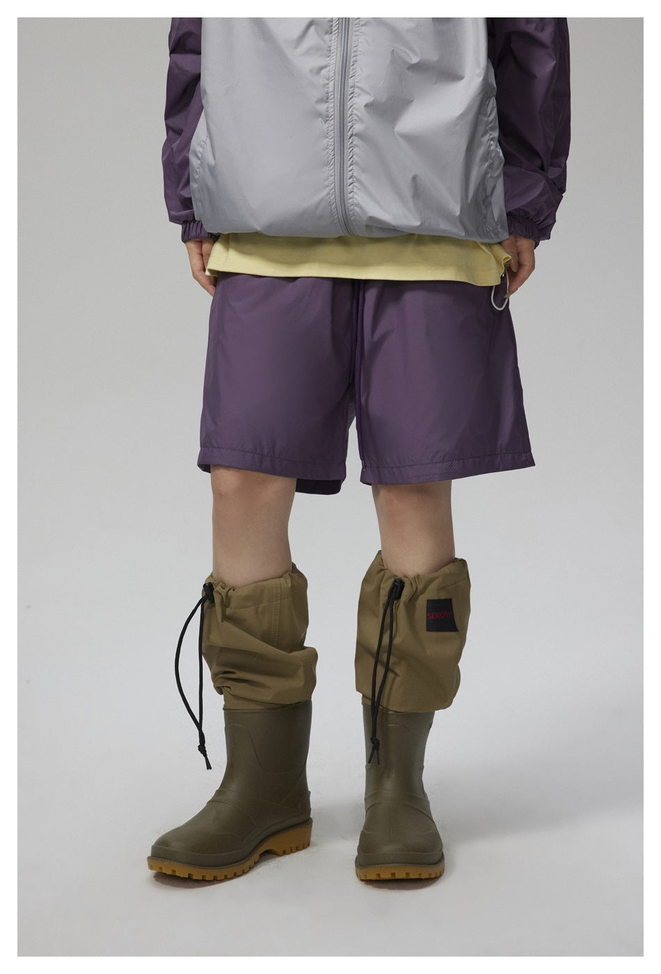 Close crop of a model wearing purple shorts with a gray and purple jacket, yellow tee and olive rain boots