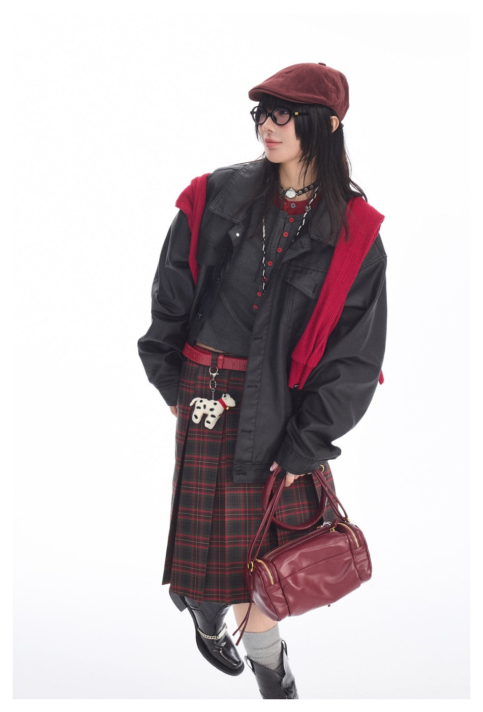 Model in a charcoal jacket, gray top, red-and-black plaid skirt, and red knit layer, carrying a burgundy bag.