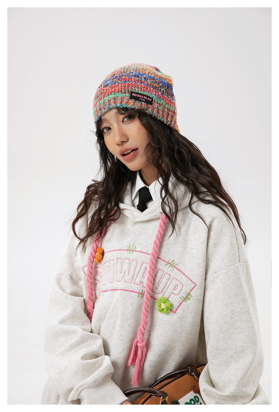 Close view of a model in a light gray hoodie with pink drawstrings and floral lettering, wearing a multicolor knit beanie.
