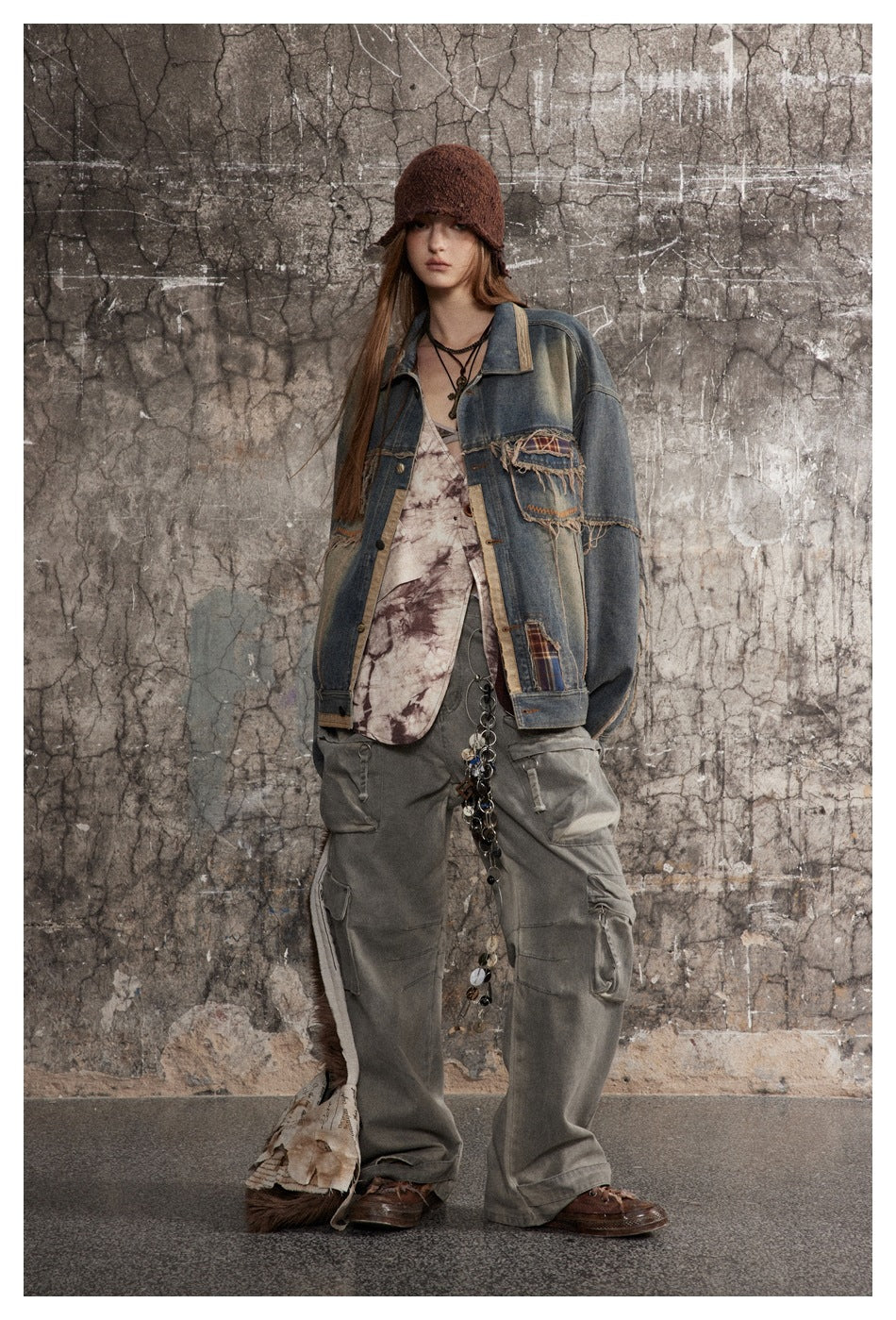 Full-length model wearing a distressed blue denim jacket with plaid patches, gray cargo pants, and a brown knit hat.