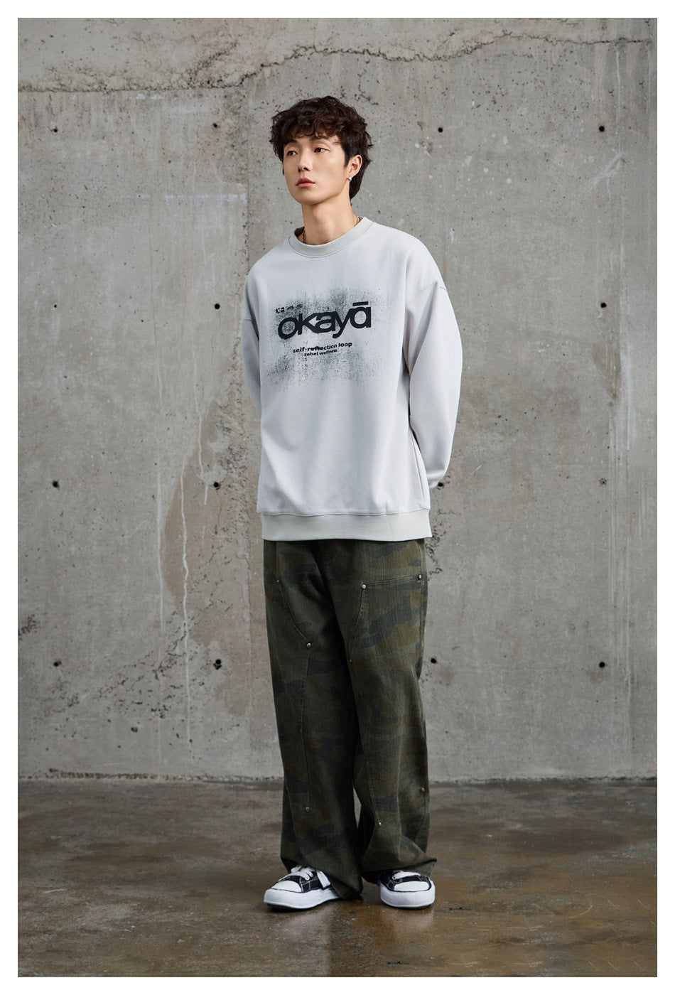 Full-length model wearing a light gray graphic sweatshirt and loose dark camouflage pants.