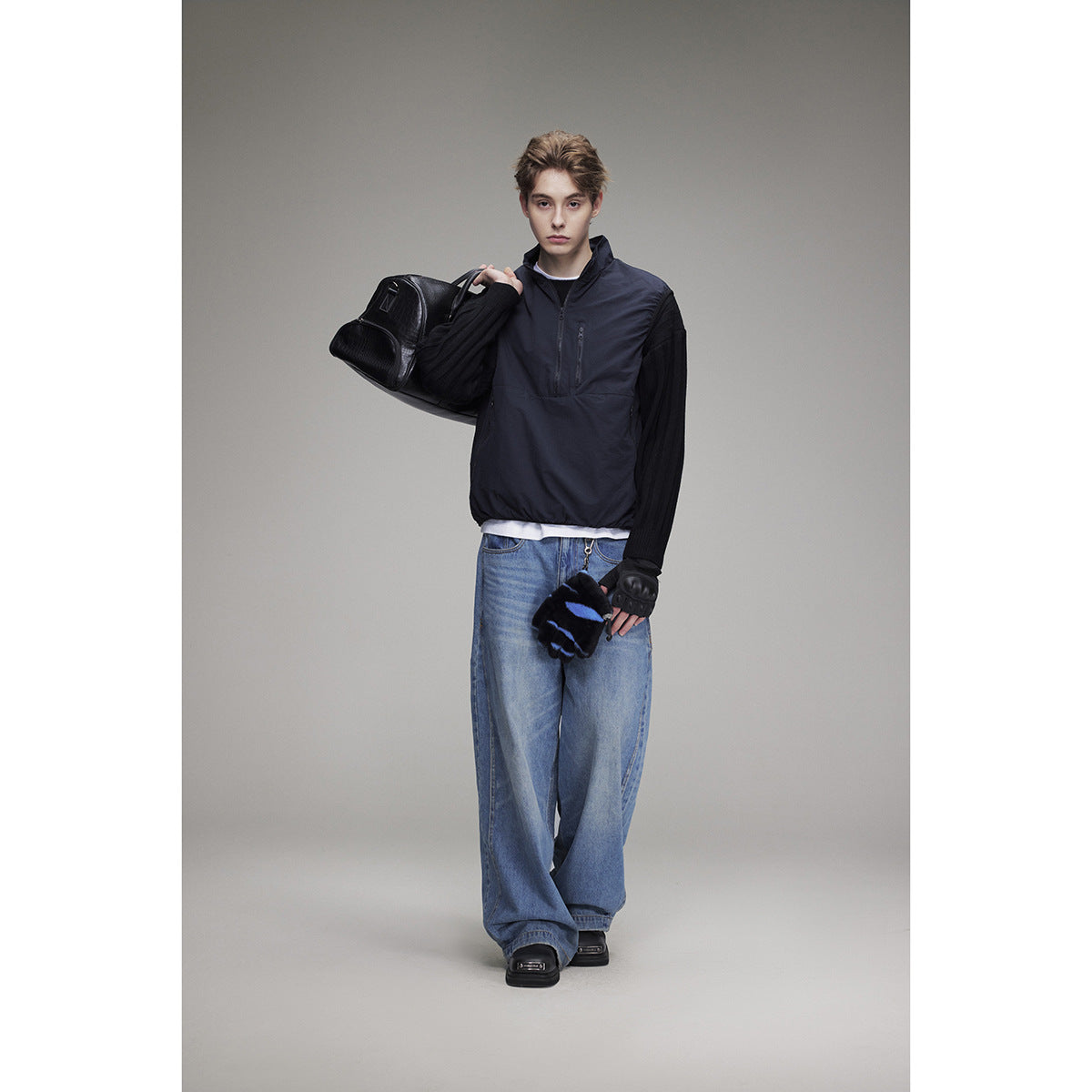 Model wearing a navy pullover vest with a quarter zip over black sleeves and loose blue jeans.