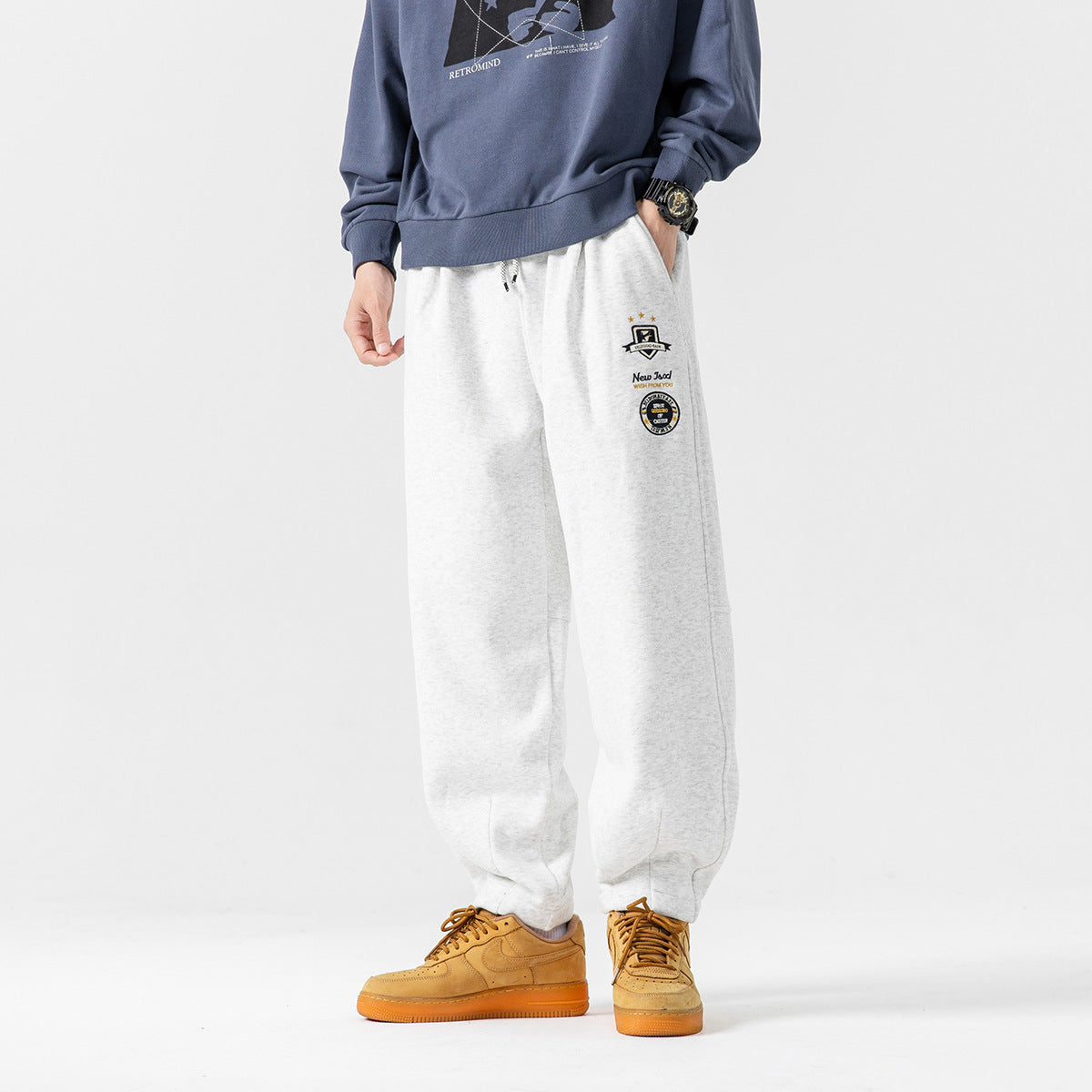 Model wearing light gray relaxed joggers with drawstring waist and small embroidered thigh patches.