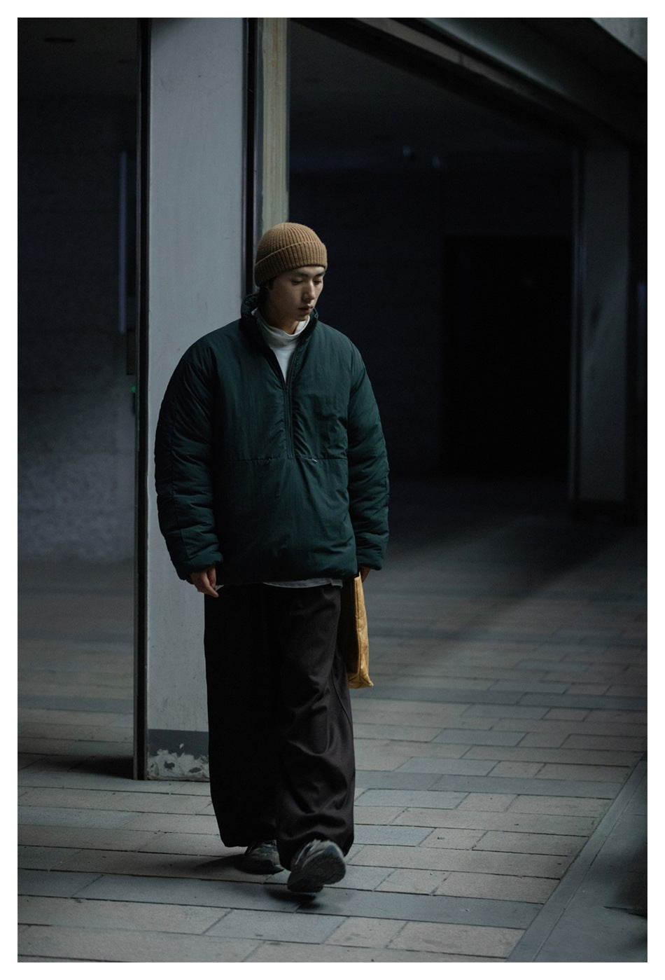 Model wearing a dark teal padded pullover jacket and tan knit beanie, shown walking outdoors.