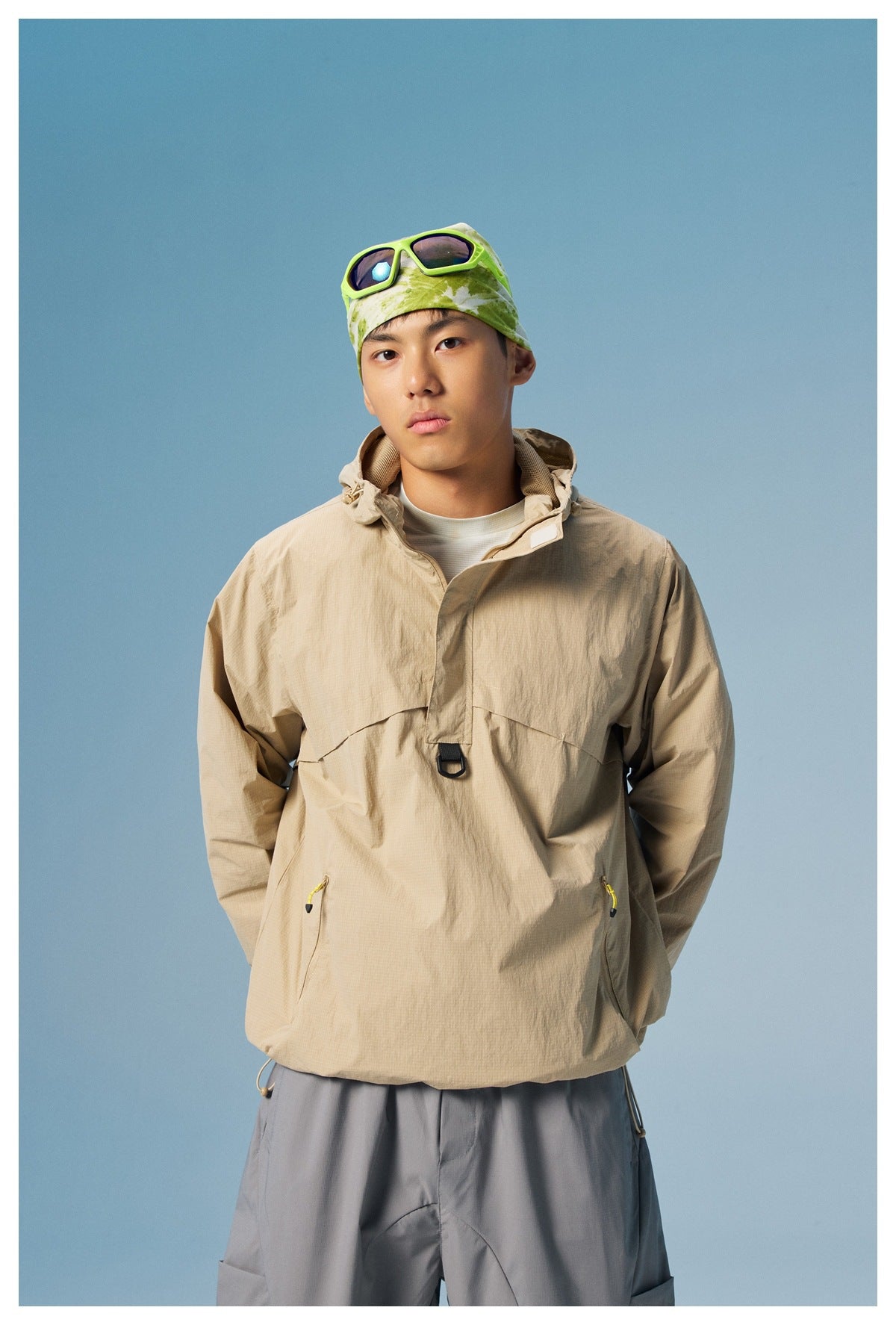 Model wearing a beige hooded pullover jacket with front pockets and gray trousers.