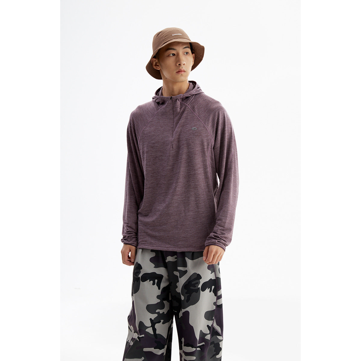 Model wearing a muted purple hooded pullover with gray-and-black camouflage pants and a brown bucket hat.