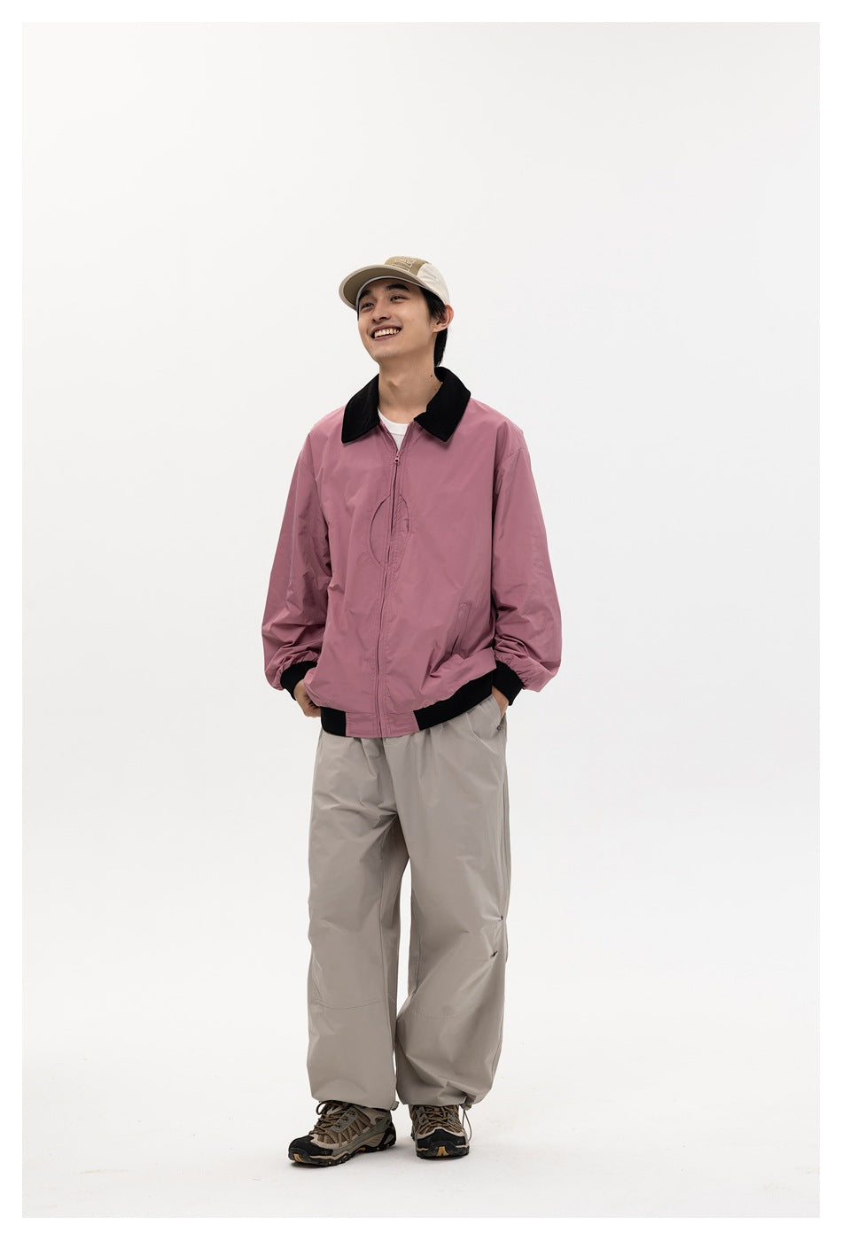 Man wearing pink zip jacket with black collar and ribbed trim, beige wide-leg pants and a cap, full-length view