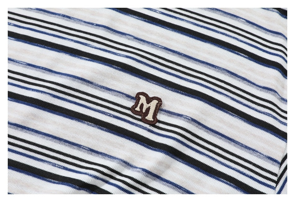 Close-up of a small brown embroidered M-shaped emblem on a cream shirt with black and blue stripes.