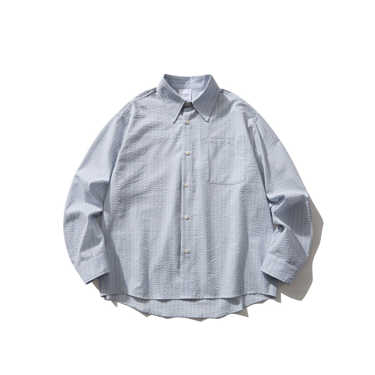 Light blue seersucker-style striped long-sleeve button-up shirt laid flat, with chest pocket and curved hem