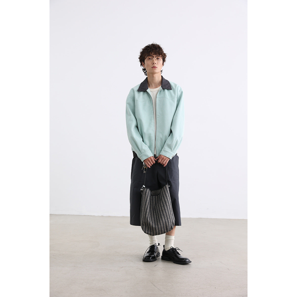 Model wearing a pale mint zip jacket with a dark collar, styled with dark shorts and a striped shoulder bag.