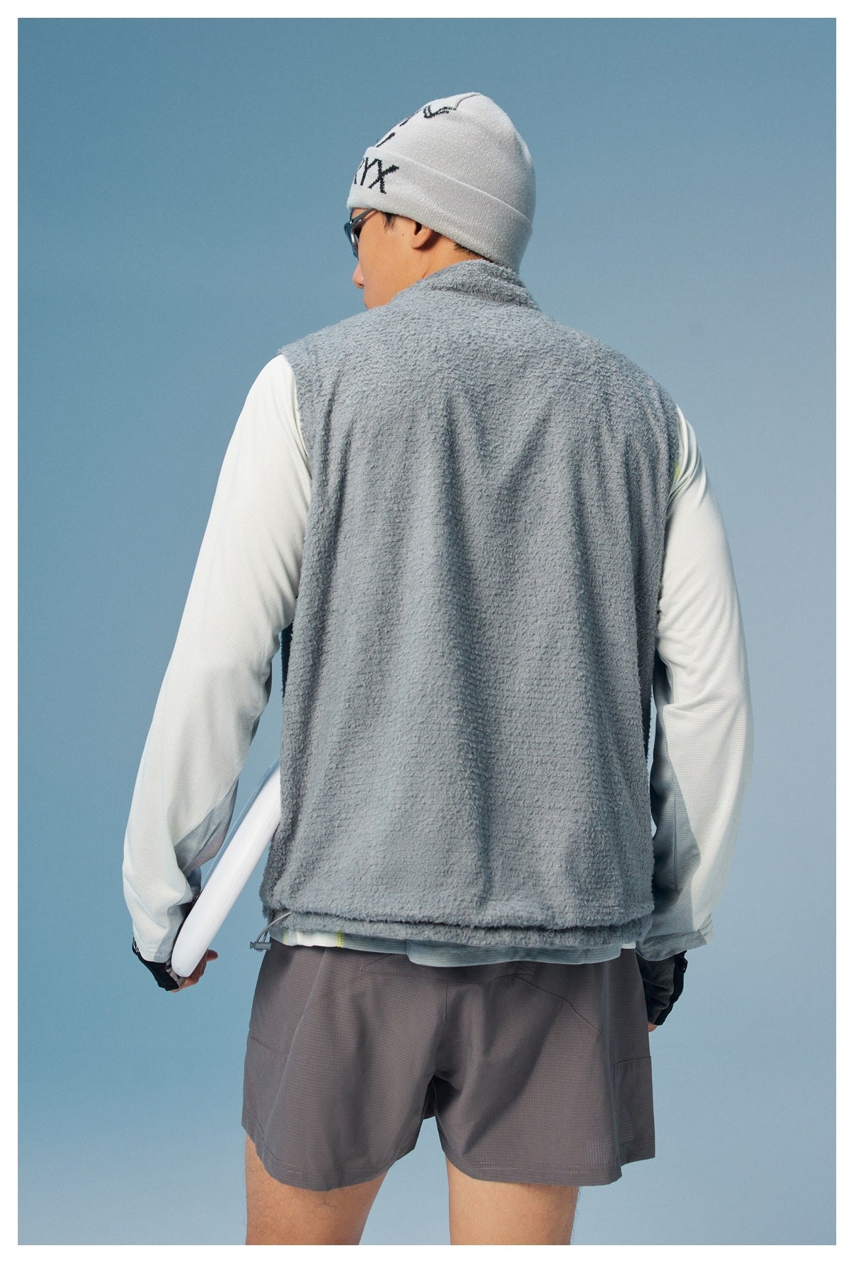 Rear view of a man wearing a gray fuzzy sleeveless vest over a pale long-sleeve top and gray shorts.