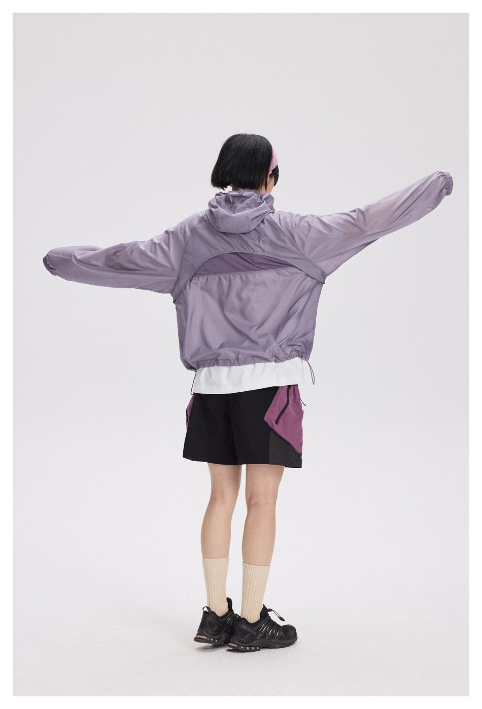 Rear view of a model wearing a lavender hooded jacket with a mesh back vent and black shorts.