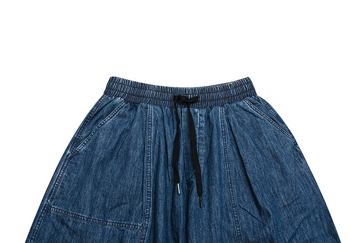 Cropped front view of blue denim trousers showing the elastic waistband, black drawstring, and large front pockets.