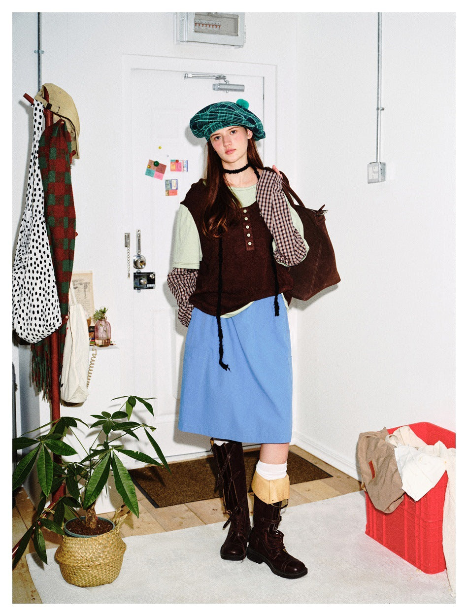 Model wearing a brown sleeveless top over layered shirts and a blue skirt, with a teal plaid beret.
