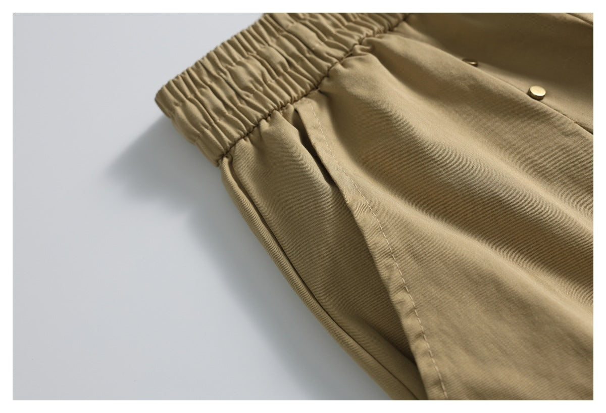 Close-up of tan shorts with a gathered elastic waistband, side pocket seam, and small gold-tone button.