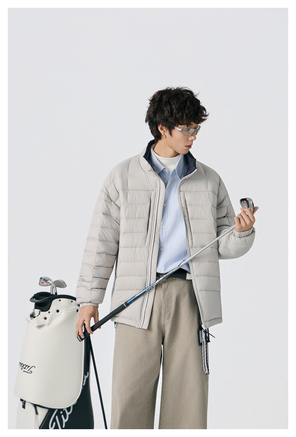 Model wearing a light gray quilted jacket with a navy-lined stand collar, holding a golf club beside a golf bag.