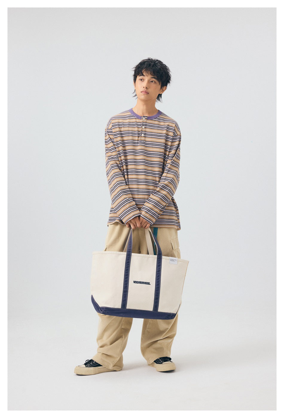Full-length model wearing a tan striped henley and beige cargo pants, holding a large cream tote with navy trim.