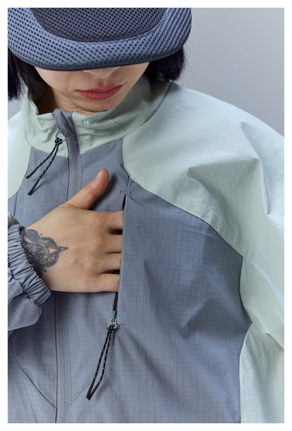 Close-up of a model wearing a slate-gray and pale sage color-block jacket with a high collar and zip pockets.