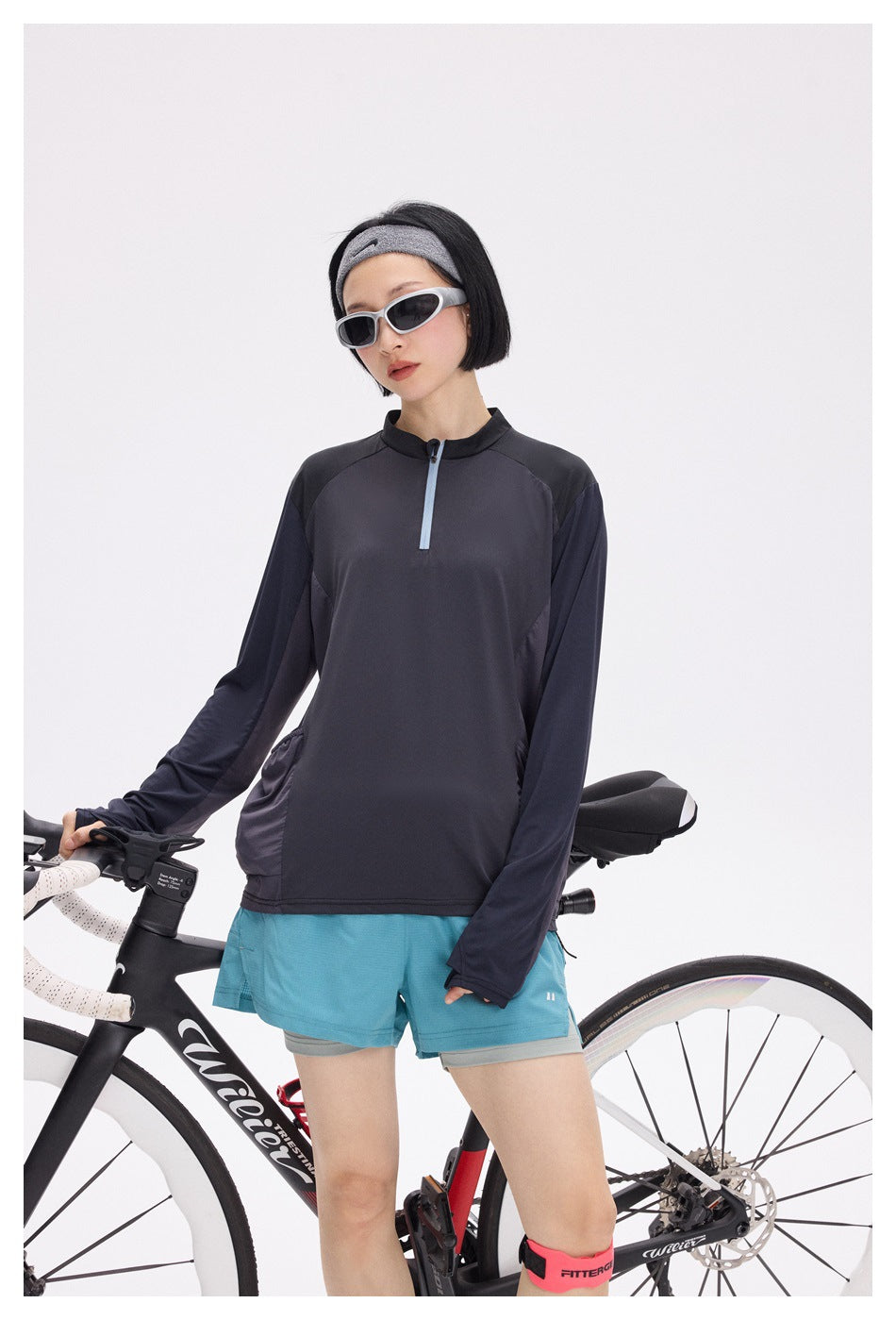 Woman wearing a charcoal and navy quarter-zip cycling top with aqua shorts, standing beside a road bike.