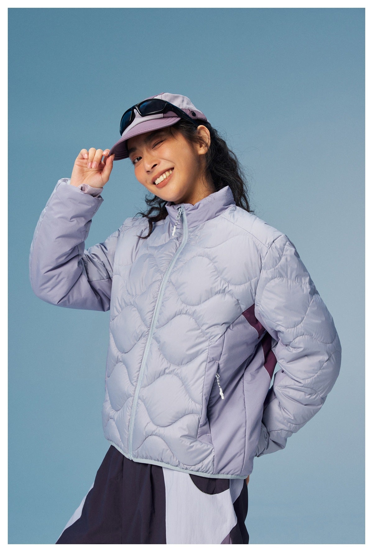 Model wears a pale lavender wave-quilted zip jacket, shown at an angle with one hand on her cap brim.